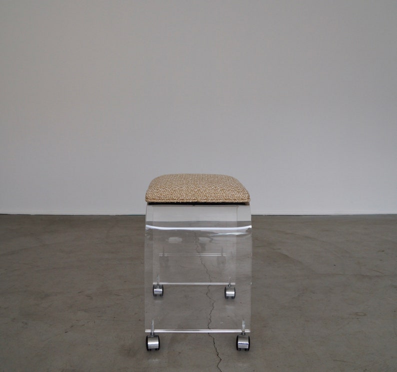 May include: A clear acrylic stool with a rectangular shape and a cushioned seat. The seat has a beige and brown patterned fabric. The stool has four small wheels for mobility. The design is modern and minimalist.
