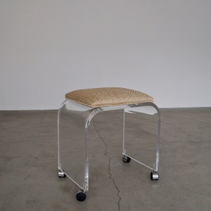May include: A clear acrylic stool with a rectangular, cushioned seat in a tan woven fabric. The stool has a curved design and sits on four small wheels.