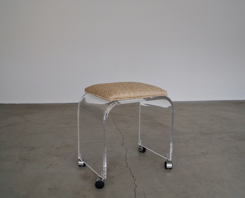May include: A clear acrylic stool with a rectangular, cushioned seat in a tan woven fabric. The stool has a curved design and sits on four small wheels.