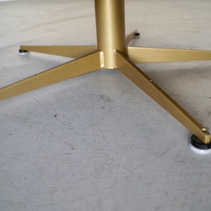 May include: A gold-colored metal table base with a central column and four angled legs. Each leg has a black and white foot pad. The base is on a gray surface.