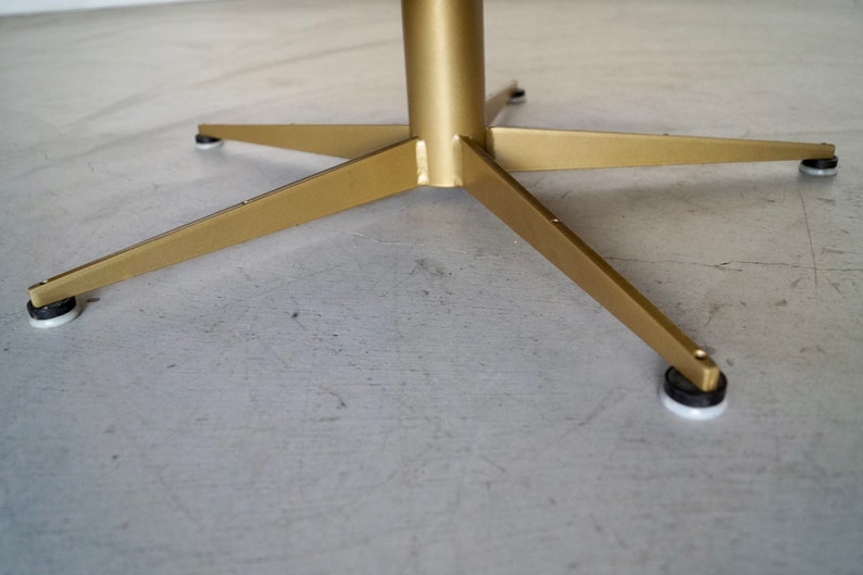May include: A gold-colored metal table base with a central column and four angled legs. Each leg has a black and white foot pad. The base is on a gray surface.
