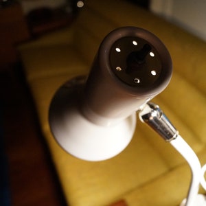 May include: A white and beige desk lamp with a round, perforated head. The lamp has a flexible arm and a circular base. The background includes a yellow sofa and a dark wooden floor.