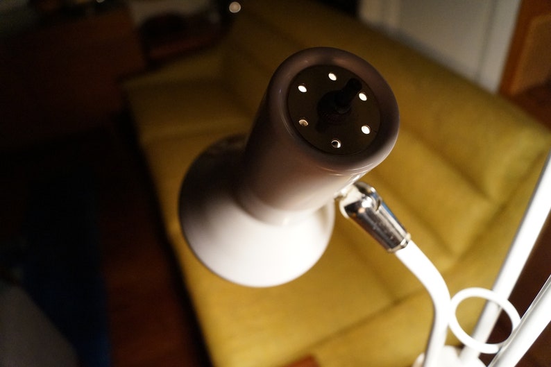May include: A white and beige desk lamp with a round, perforated head. The lamp has a flexible arm and a circular base. The background includes a yellow sofa and a dark wooden floor.