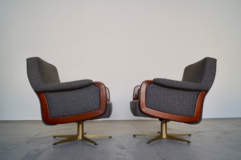 May include: Two gray upholstered swivel chairs with wooden trim and brass-colored bases. The chairs have a mid-century modern design, with curved armrests and backs. The chairs are set against a plain white wall.