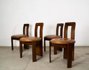 May include: Four wooden chairs with brown seats and dark brown frames. The chairs have a simple, geometric design with a rounded seat and a rectangular backrest. The chairs are arranged in a row against a white wall.