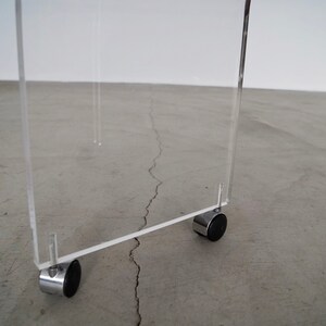 May include: A clear acrylic table with a rectangular design. The table has four chrome legs with small black wheels. The table is designed for mobility and is suitable for various uses.