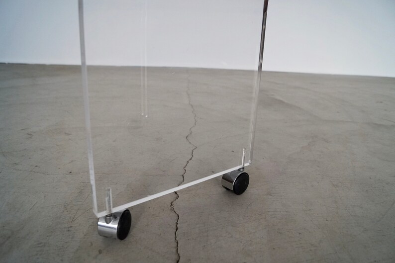 May include: A clear acrylic table with a rectangular design. The table has four chrome legs with small black wheels. The table is designed for mobility and is suitable for various uses.