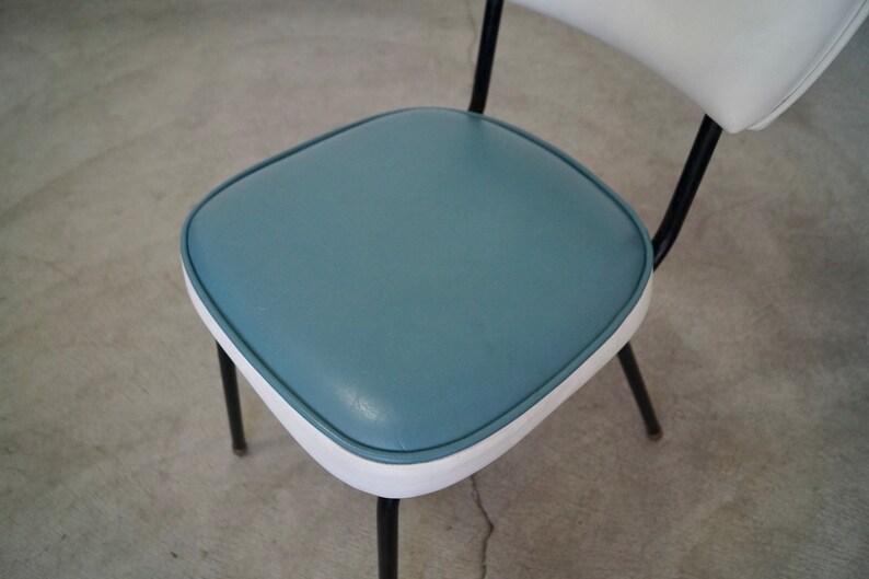 May include: A retro-style chair with a blue seat and white backrest. The seat is square with rounded corners and a white trim. The chair has black metal legs and a curved back support. The design is reminiscent of mid-century modern furniture.