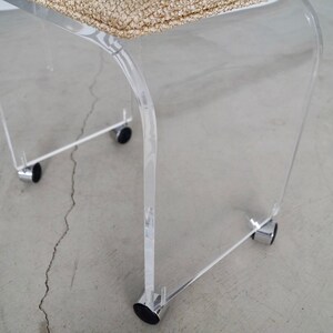 May include: A clear acrylic chair with a woven beige seat. The chair has a curved design and sits on four small black wheels attached to chrome supports. The chair is designed for indoor use.