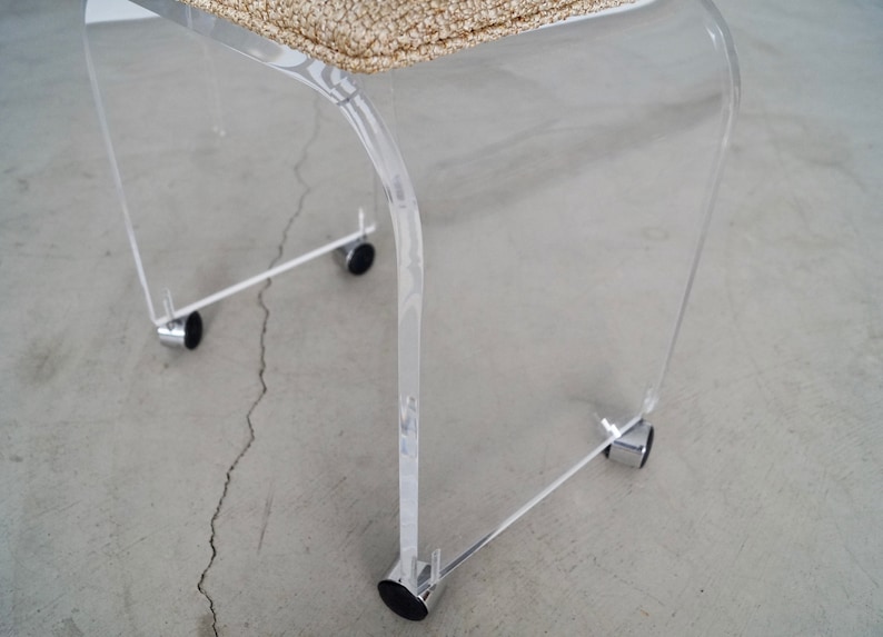 May include: A clear acrylic chair with a woven beige seat. The chair has a curved design and sits on four small black wheels attached to chrome supports. The chair is designed for indoor use.