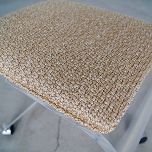 May include: A clear acrylic stool with a square, beige textured seat. The seat has a woven appearance. The stool has a modern design, with a curved base and chrome accents on the feet.