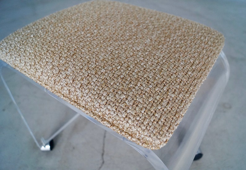 May include: A clear acrylic stool with a square, beige textured seat. The seat has a woven appearance. The stool has a modern design, with a curved base and chrome accents on the feet.