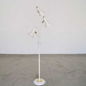 May include: A white, mid-century modern floor lamp with three adjustable spotlights. The lamp has a slender, vertical pole and a circular base with a gold-colored accent. The spotlights are angled upwards, and the lamp is set against a plain white background.