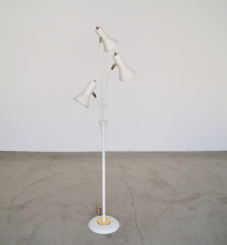 May include: A white, mid-century modern floor lamp with three adjustable spotlights. The lamp has a slender, vertical pole and a circular base with a gold-colored accent. The spotlights are angled upwards, and the lamp is set against a plain white background.