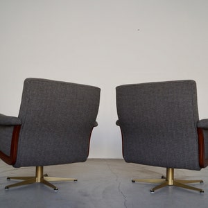 May include: Two gray upholstered swivel chairs with wooden accents and brass-colored star-shaped bases. The chairs have curved armrests and high backs, set against a plain white wall.