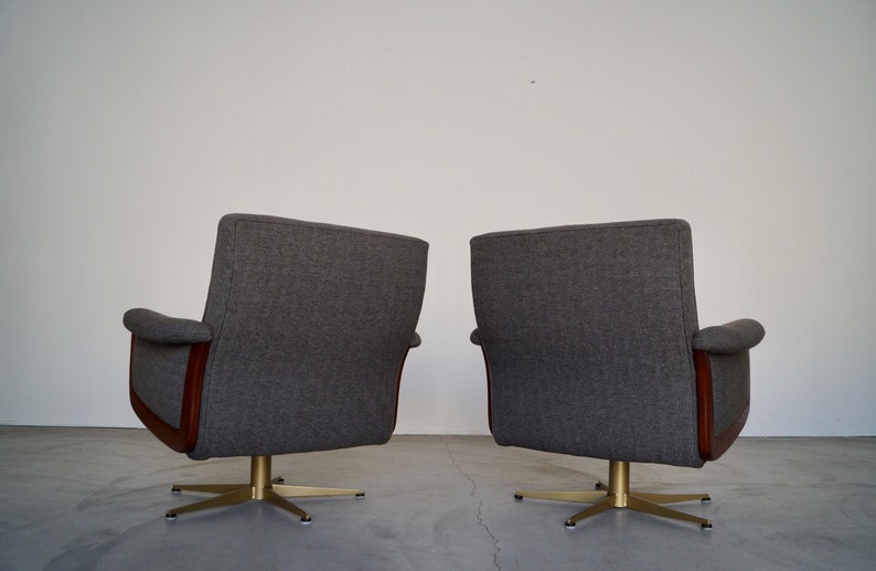 May include: Two gray upholstered swivel chairs with wooden accents and brass-colored star-shaped bases. The chairs have curved armrests and high backs, set against a plain white wall.