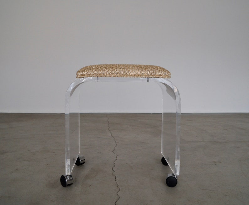 May include: A clear acrylic stool with a rectangular, textured, beige seat. The stool has a curved design and sits on four black and chrome casters for easy mobility. The modern design is suitable for various interior styles.
