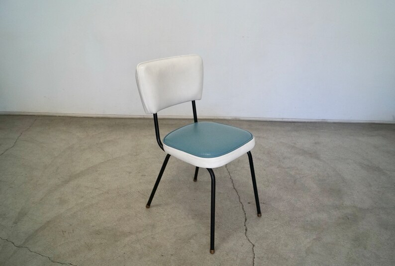 May include: A retro-style chair with a white backrest and a teal seat, outlined in white. The chair has black metal legs with brass-colored tips. The chair is a vintage piece, perfect for a kitchen or dining room.