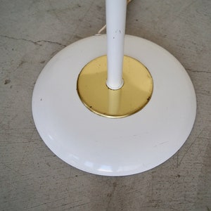 May include: A white floor lamp base with a gold-colored center. The base is round and has a white stem. The lamp is on a gray concrete floor.