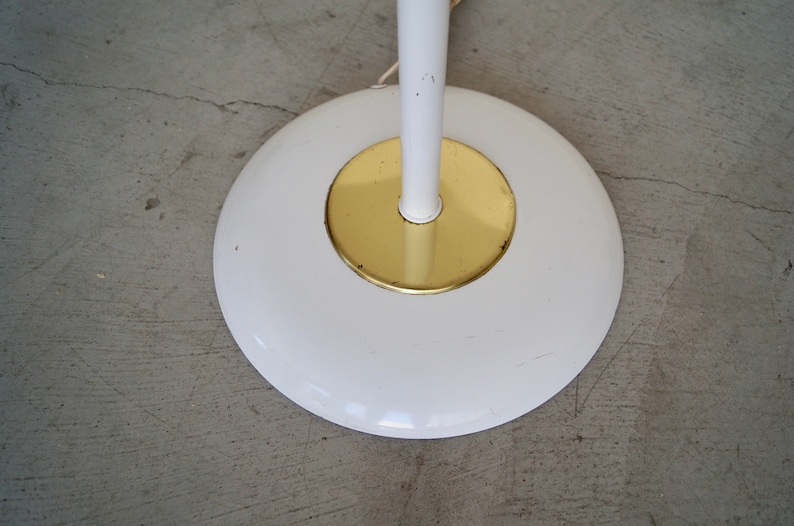 May include: A white floor lamp base with a gold-colored center. The base is round and has a white stem. The lamp is on a gray concrete floor.