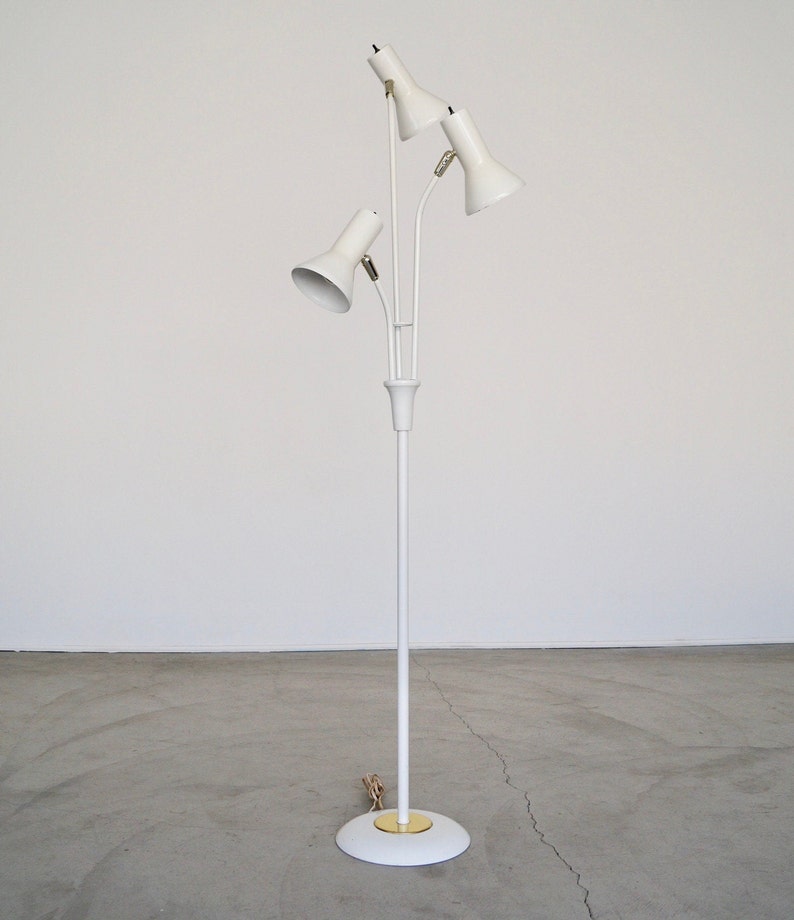 May include: A white, mid-century modern floor lamp with three adjustable spotlights. The lamp has a slender, white pole and a circular base with a gold-colored accent. The spotlights are angled and directed upwards.