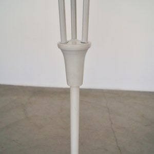 May include: A white, minimalist floor lamp with a slender, vertical pole and a decorative top featuring three upward-curving supports. The lamp's design is simple and modern, suitable for various interior styles.