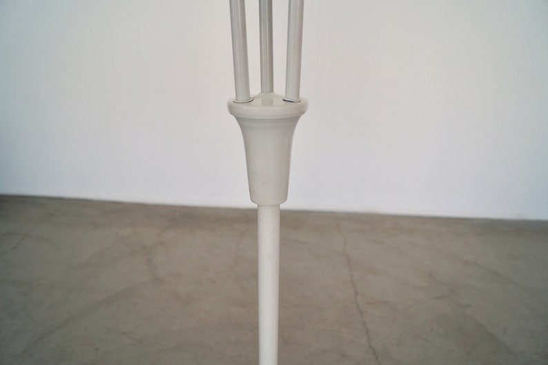 May include: A white, minimalist floor lamp with a slender, vertical pole and a decorative top featuring three upward-curving supports. The lamp's design is simple and modern, suitable for various interior styles.
