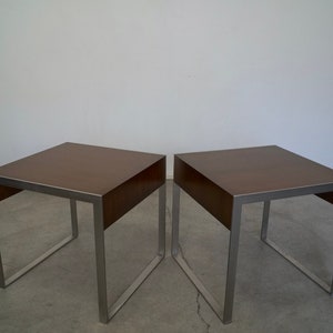 May include: Two square side tables with brown tops and silver metal frames. The tables have a minimalist design, with a simple, clean aesthetic. The tables are suitable for a living room or bedroom.
