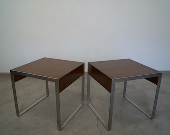 May include: Two square side tables with brown tops and silver metal frames. The tables have a minimalist design, with a simple, clean aesthetic. The tables are suitable for a living room or bedroom.