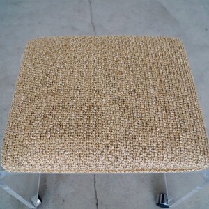 May include: A square, low-profile stool with a woven, beige fabric seat. The legs are clear acrylic, and the stool has small, silver-colored casters for mobility. The design is modern and minimalist.