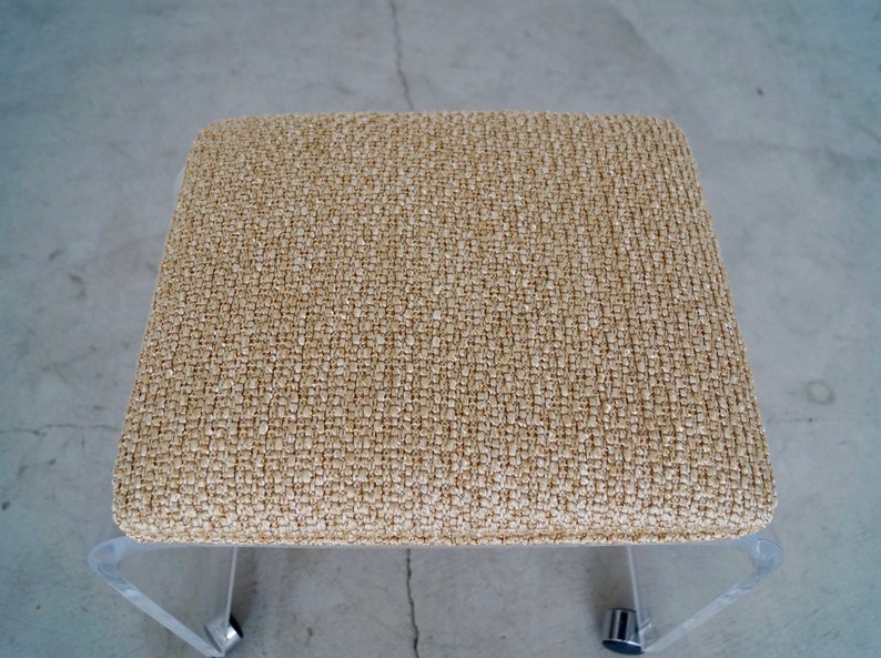 May include: A square, low-profile stool with a woven, beige fabric seat. The legs are clear acrylic, and the stool has small, silver-colored casters for mobility. The design is modern and minimalist.