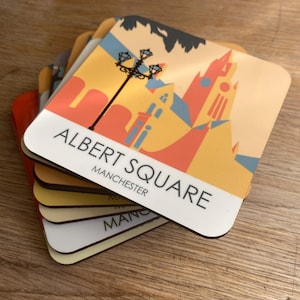 May include: A stack of six colourful coasters featuring a retro-style illustration of Albert Square in Manchester, England. The top coaster has the text "ALBERT SQUARE" and "MANCHESTER" printed on it.