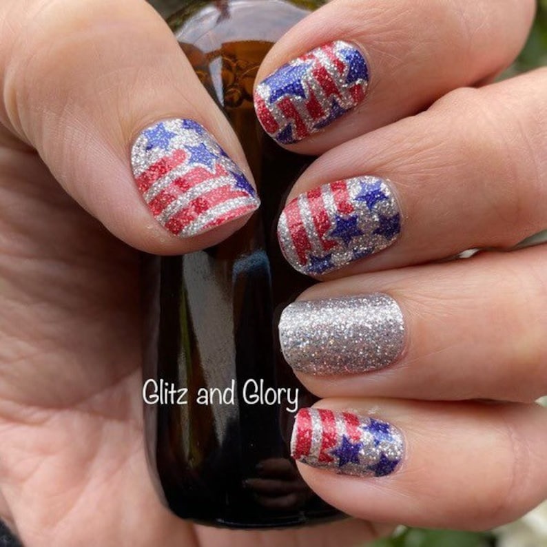 May include: Close-up of a hand with red, white and blue glitter nail polish with stars and stripes design. The text "Glitz and Glory" is visible on the bottle.