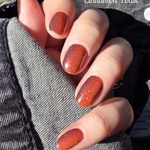 May include: Close-up of a hand with polished nails. The nails are painted in a gradient of orange and brown with a glittery finish. The text "Cinnamon Toast" is visible in the upper right corner.