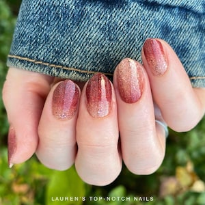 May include: A hand with polished nails in a gradient of red and gold glitter. The nails transition from a deep red at the base to a shimmering gold at the tips. The text "LAUREN'S TOP-NOTCH NAILS" is at the bottom.