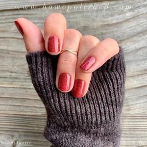 May include: Close-up of a hand with red glitter nail polish. The nails are painted with a vibrant, sparkling red color. A gold ring is worn on the ring finger. The hand is covered by a dark gray knitted sleeve. The website address www.howepolished.com and the word "cran-tastic" are visible.