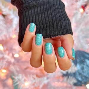 May include: Close-up of a hand with teal glitter nail polish. The nails are short and rounded. The hand is wearing a black sweater.