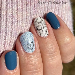 May include: A hand with manicured nails. The nails feature a variety of designs, including solid teal polish, a white base with black heart doodles, and a light blue glitter with a black heart. The background is a light pink knitted fabric.