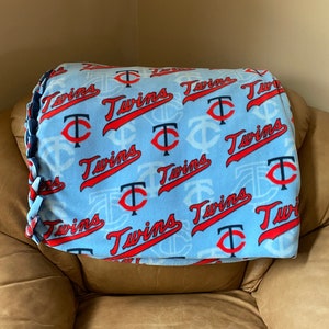 May include: A blue fleece blanket with a repeating pattern of the Minnesota Twins logo. The logo is red and white with a black outline. The blanket has a braided edge.