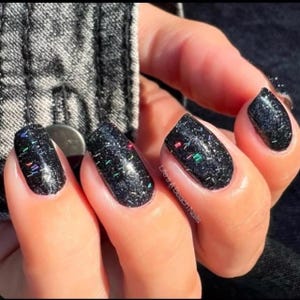 May include: Close-up of a hand with manicured nails. The nails are painted black with a glittery, speckled finish, featuring colorful flecks. The text "Life's A Beach Nails" is visible on one nail.