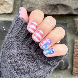 May include: Close-up of a hand with red, white and blue nail art featuring American flag designs. The nail art is called "Patriotic Parade".