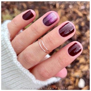 May include: A set of two thin, smooth rose gold rings worn on a finger. The fingernails are painted with a glossy deep burgundy polish, with one nail featuring a glittery purple and silver ombre gradient. Text reads: Vintage Velvet + Rosé Ombré.
