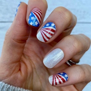 May include: Close-up of a hand with red, white and blue nail polish featuring an American flag design. The nails are decorated with a glitter polish and a white base.