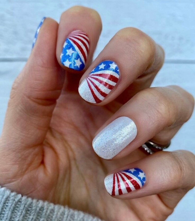 May include: Close-up of a hand with red, white and blue nail polish featuring an American flag design. The nails are decorated with a glitter polish and a white base.