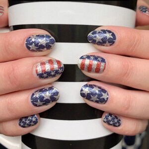 May include: Close-up of hands with red, white and blue glitter nail polish. The nails are decorated with stars and stripes.