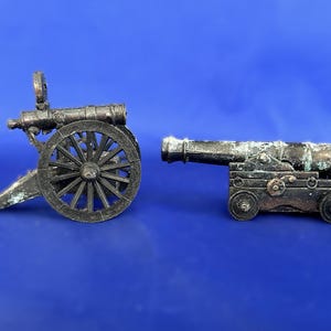 May include: Two miniature metal cannons with a weathered, dark metallic finish. One cannon features large spoked wheels and a trailing support, while the other is mounted on a rectangular carriage with four small wheels and visible side detailing.