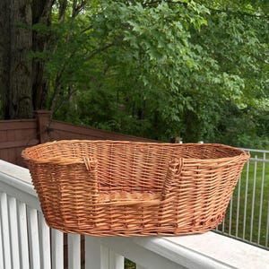 May include: An oval-shaped pet bed constructed from woven light brown wicker. The basket features a low-profile front opening for easy access, with a sturdy, continuous rim and a flat, woven base, showcasing a natural wood-toned finish throughout.