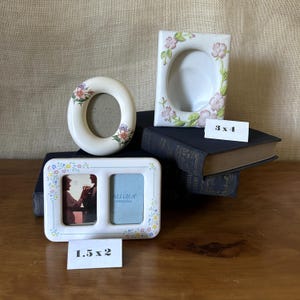 May include: Three white ceramic picture frames with floral accents. One oval frame, one rectangular frame with pink flowers, and one double-window rectangular frame with small colorful flowers. Labels indicate sizes 3 x 4 inches and 1.5 x 2 inches.