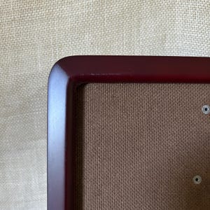 May include: A close-up view of the corner of a rectangular picture frame with a smooth, dark burgundy finish. The frame surrounds a textured, medium-brown fabric insert. Two small, circular metal grommets are visible on the right side of the insert.