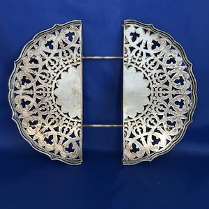 May include: Silver-colored, ornate trivet with a filigree design. The trivet is made of two semi-circular halves connected by a metal bar. The edges are scalloped, and the design features intricate patterns. The background is a solid blue.